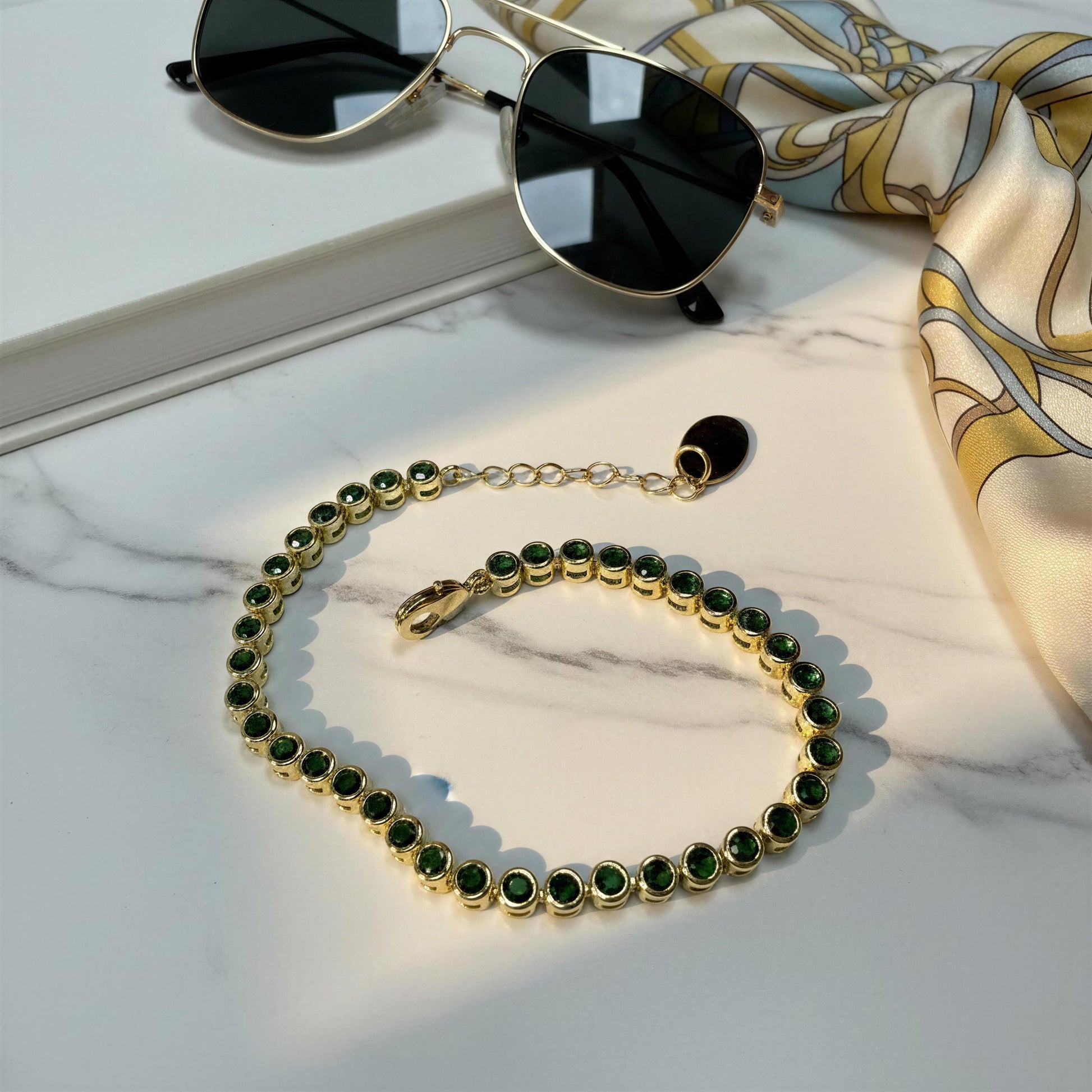 The Siena Emerald Tennis Bracelet displayed on a white marble surface next to sunglasses and a patterned silk scarf.