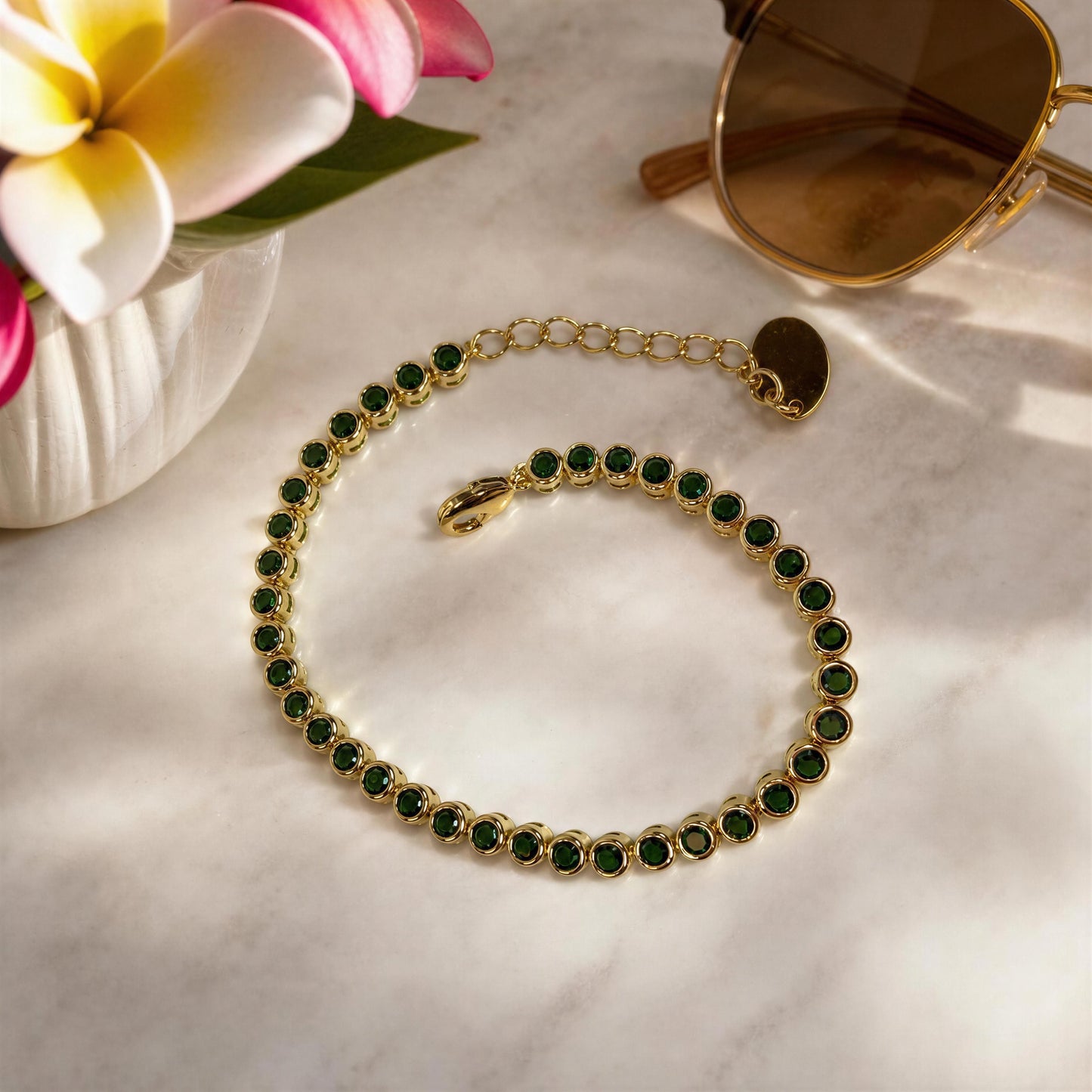 The Siena Emerald Tennis Bracelet, from Chica Chica Jewelry, arranged in a spiral on a light marble surface with decorative flowers and sunglasses nearby.