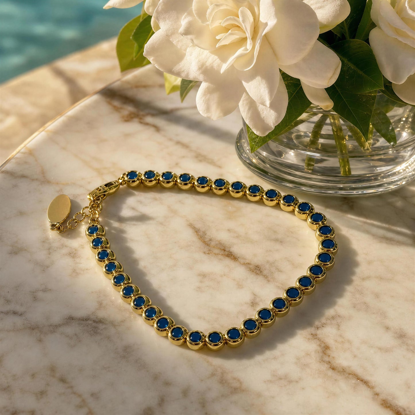 An 18k gold plated bracelet with turquoise gemstones, arranged on a light marble surface beside white flowers in a glass vase.