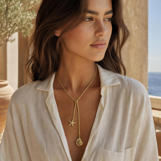 A woman wearing the Sirena Lariat Necklace from  Chica Chica Jewelry, a delicate gold lariat necklace with a starfish and seashell charms, styled with a white linen shirt in a warm, sunlit outdoor setting.