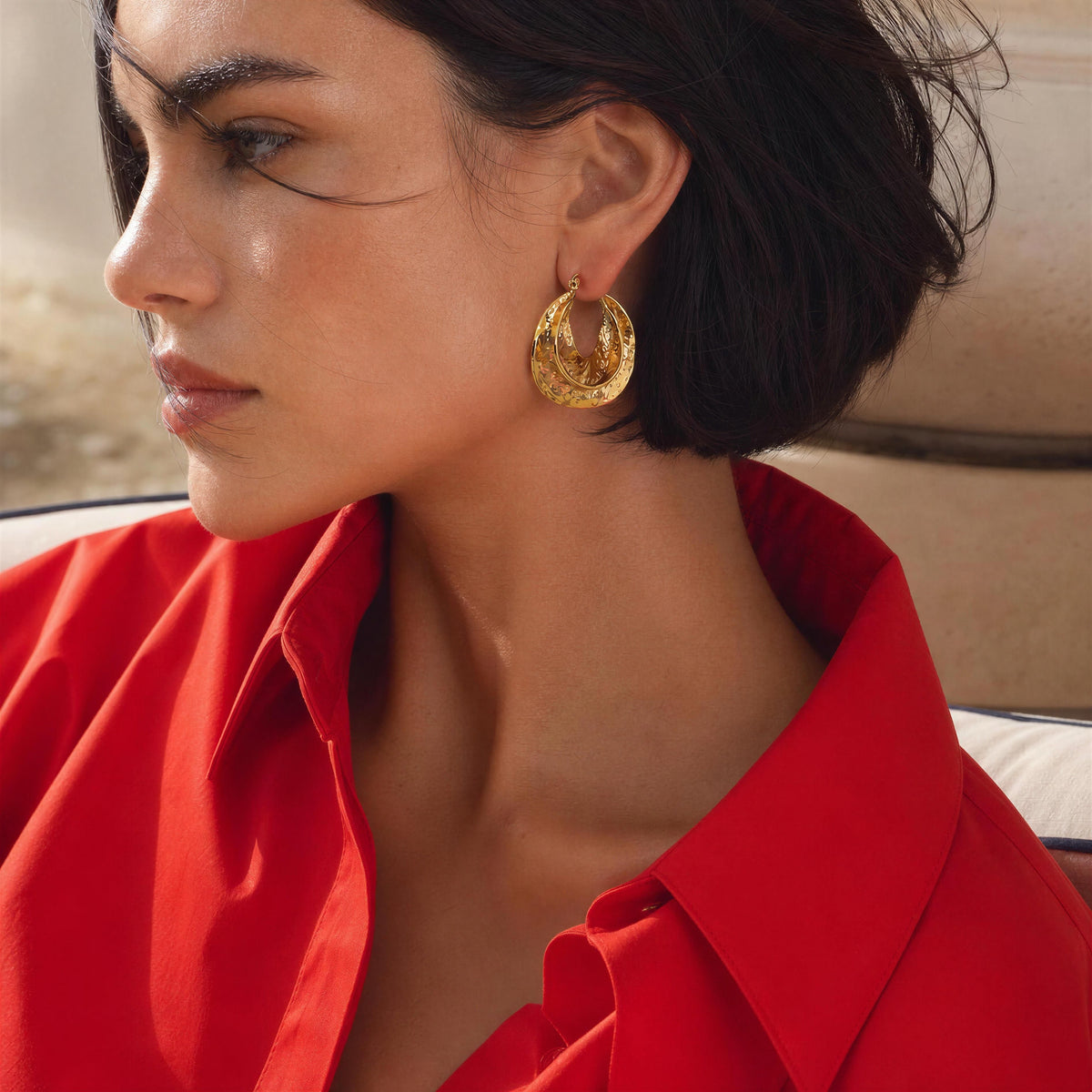 A close-up side profile of a woman with short dark hair wearing the Solea Earrings from Chica Chica Jewelry, a textured gold huggie hoop earring and a bright red collared shirt.