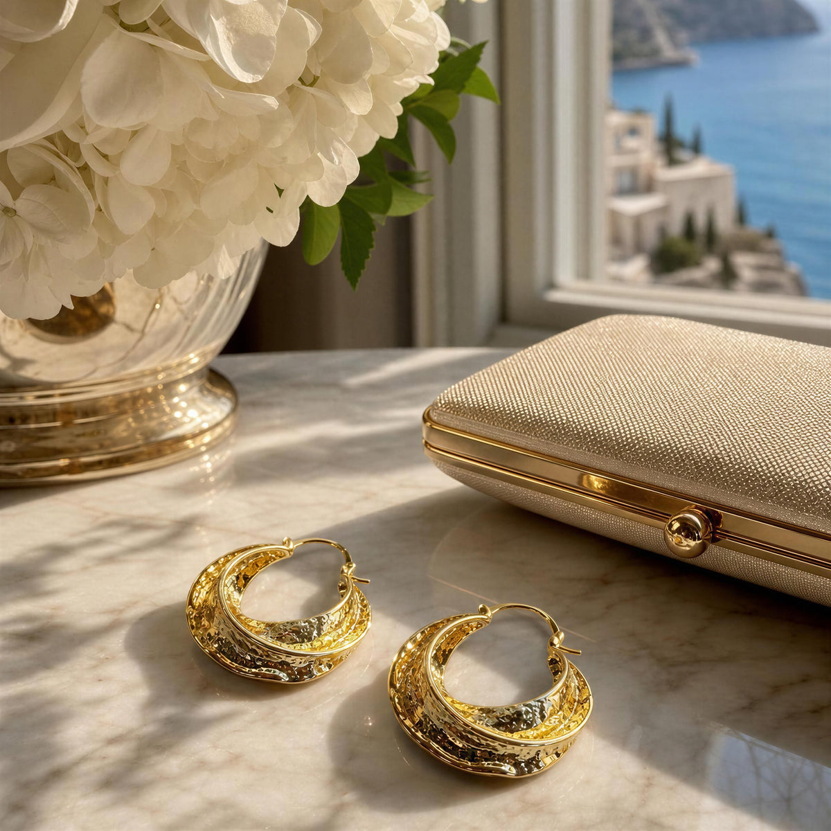 Two textured gold crescent hoop earrings displayed on a marble surface next to a gold clutch case, with a vase of white flowers and a scenic window view in the background.