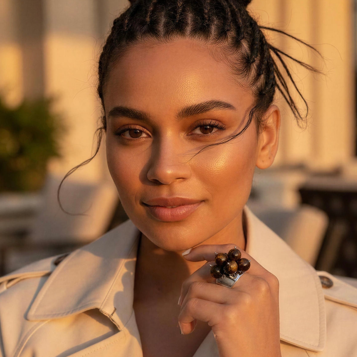 A close-up portrait of a woman with braided hair wearing a cream trench coat and the Talia Ring from Chica Chica Jewelry, a chunky ring with dark brown beads, set against a warm, softly blurred outdoor background.