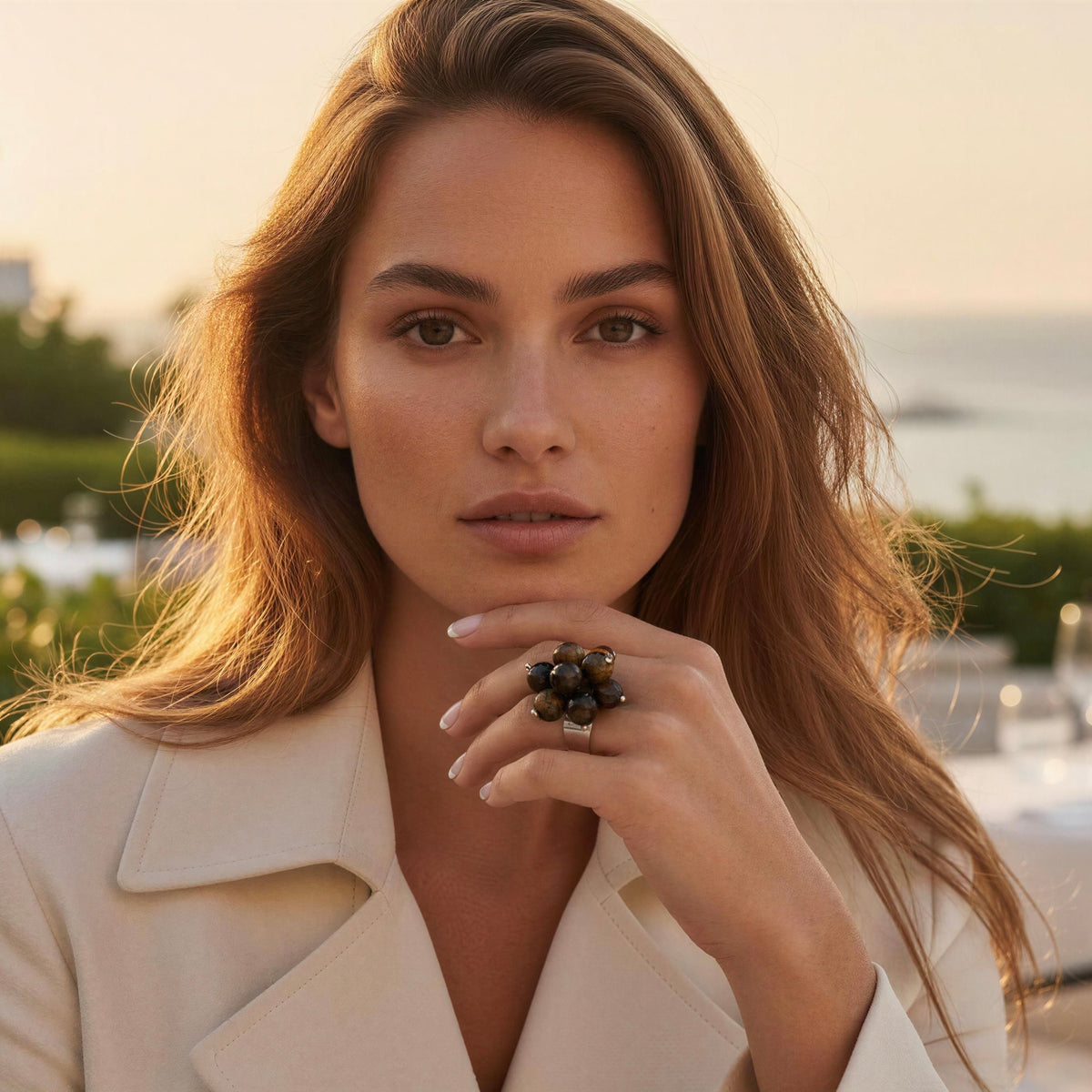 A woman in a cream-colored blazer wearing the  Talia Ring from Chica Chica Jewelry,  a large statement ring with dark, clustered stones, photographed outdoors with a warm sunset background.