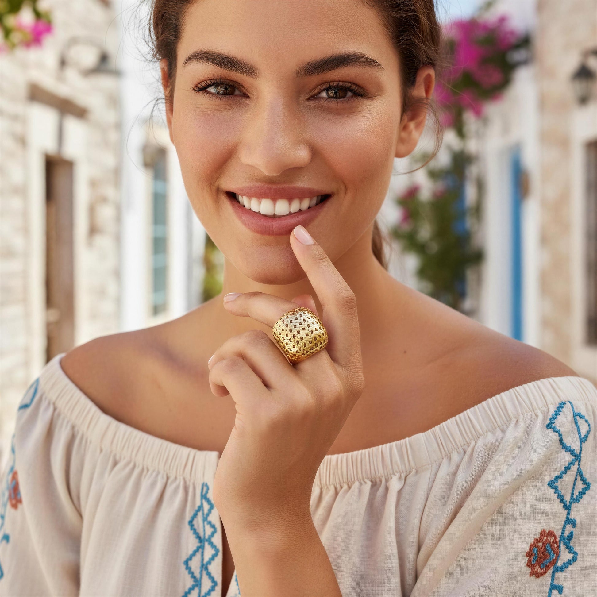 A smiling woman wearing a large textured gold ring on her finger, posing in an off-the-shoulder cream blouse with blue and orange embroidery, set against a blurred background of a European-style street.