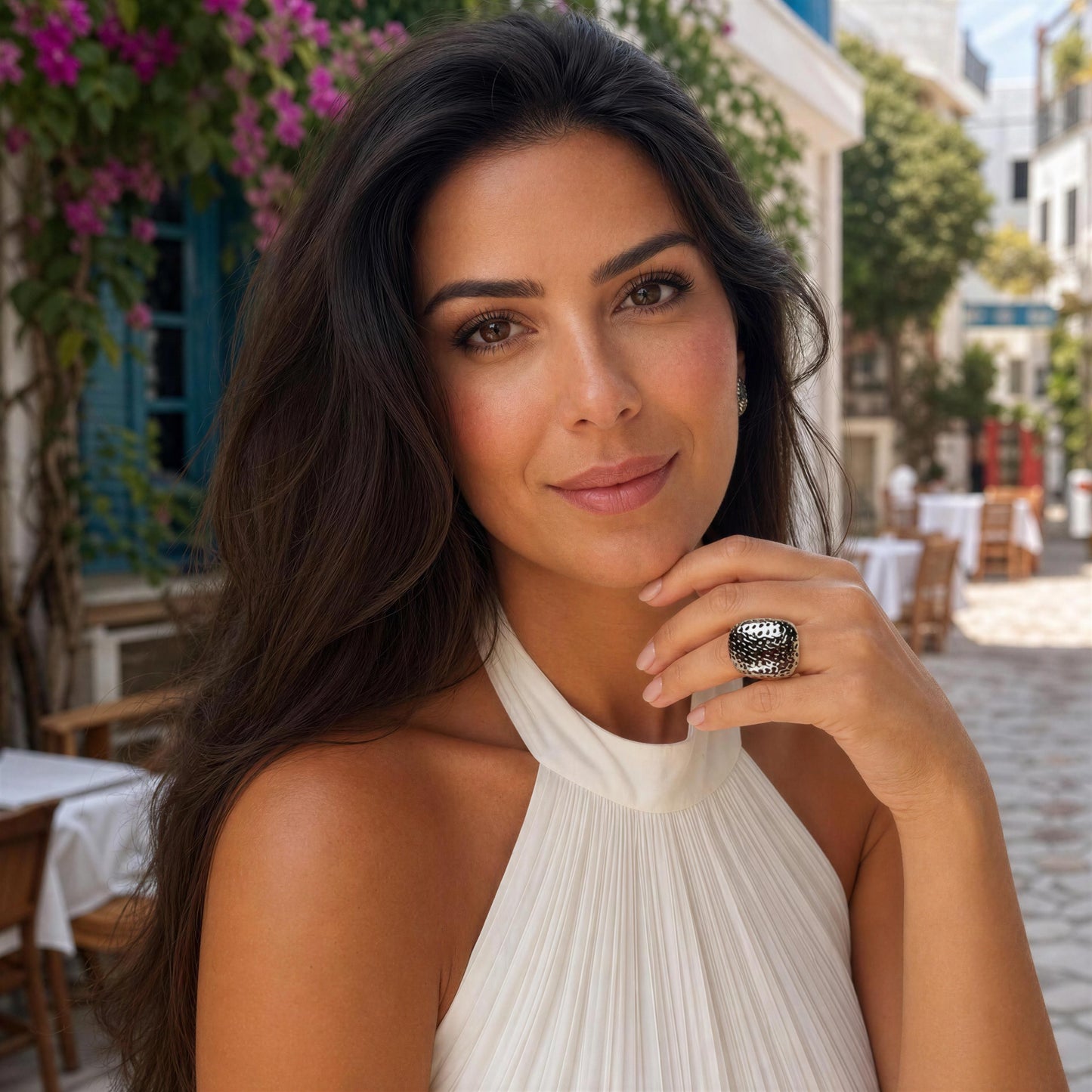 A woman with long dark hair wearing a white halter top and a large textured silver ring, posing outdoors in a sunny, Mediterranean-style street setting with flowers and tables in the background.