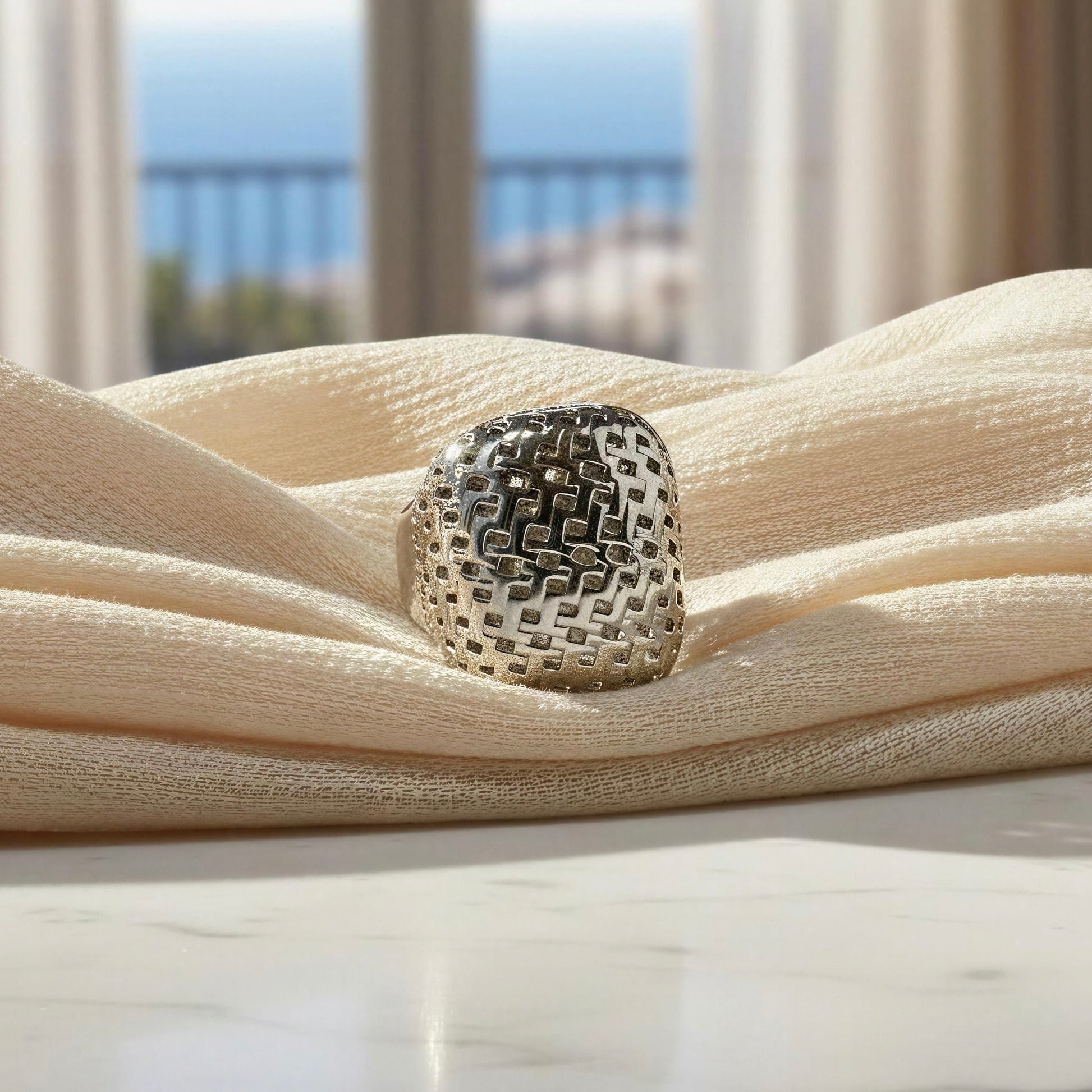 A silver ring with a textured, woven geometric pattern, resting on draped cream-colored fabric in front of a softly blurred window view.
