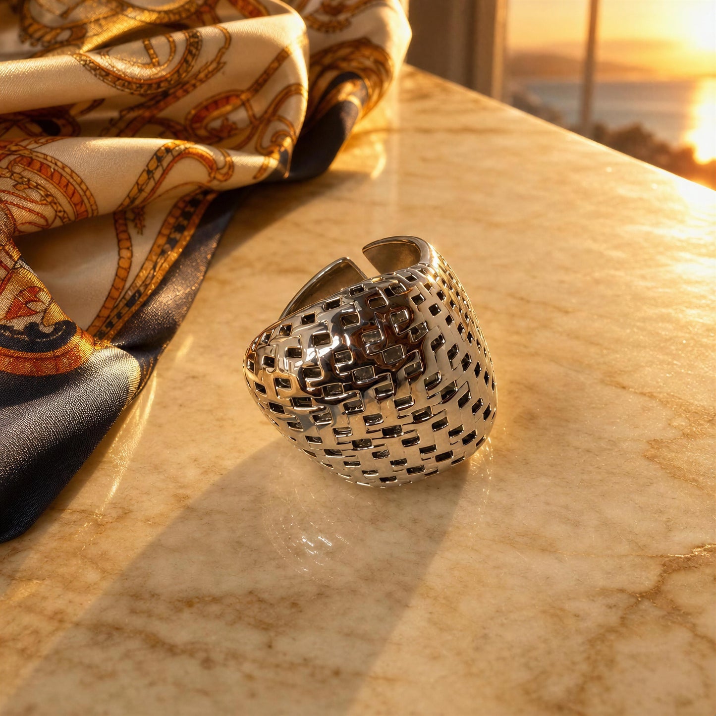 A wide silver cuff ring with a geometric, woven-texture open design, resting on a beige marble surface with a patterned silk scarf and a warm sunset background.