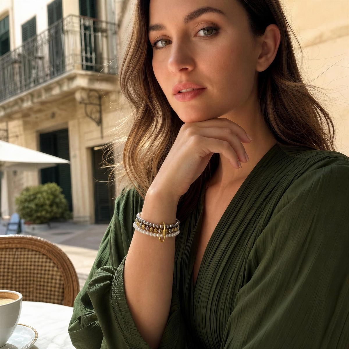 A woman wearing the Trio Radiance Bracelet a delicate multi-strand bracelet with white pearls, gold-toned beads, sitting at an outdoor cafe table.