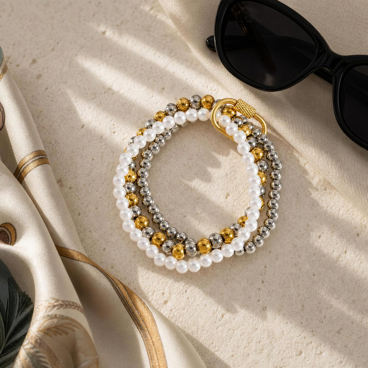 A set of three beaded stretch bracelets with white faux pearls, silver beads, and gold metallic beads, styled on a textured beige surface with black sunglasses and a cream silk scarf.