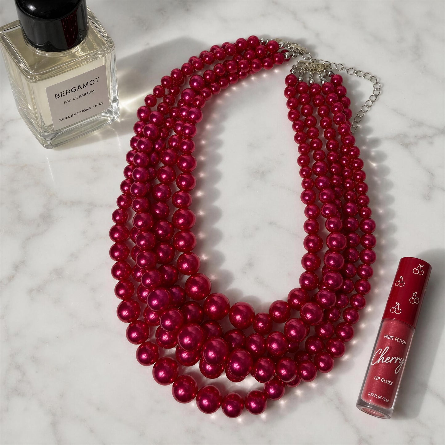 The Vienna Rose Pearl Cascade Necklace with a bottle of Bergamot perfume and Cherry lip gloss on a marble surface