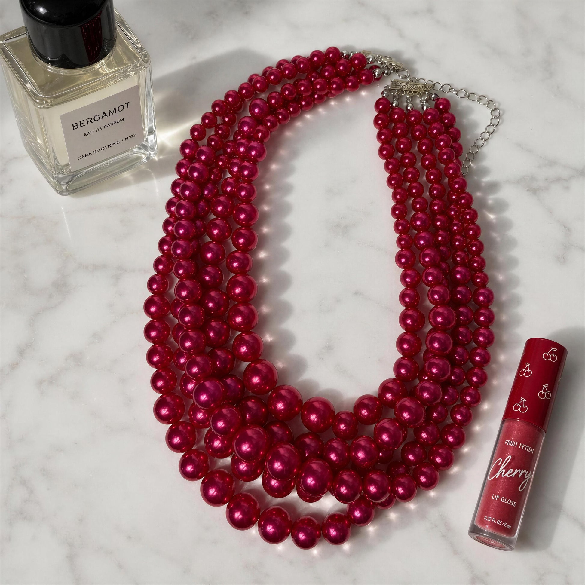 The Vienna Rose Pearl Cascade Necklace with a bottle of Bergamot perfume and Cherry lip gloss on a marble surface