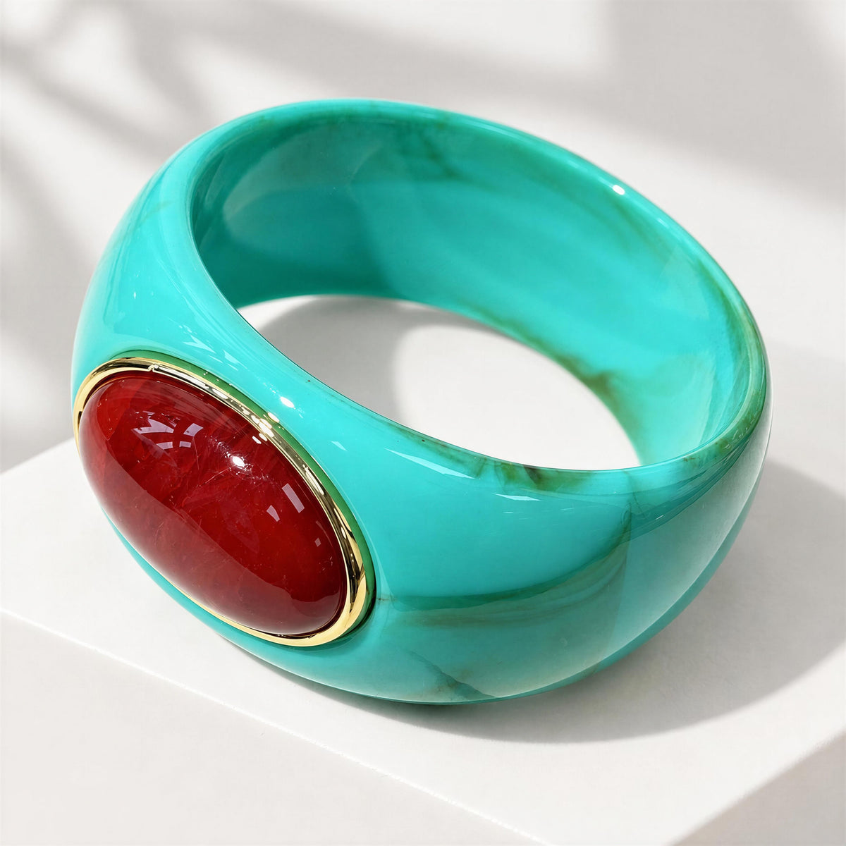 The Vittoria Bangle from Chica Chica Jewelry. A wide turquoise bangle bracelet with a glossy burgundy wine oval stone set in a gold bezel, photographed on a clean white surface with soft shadows.