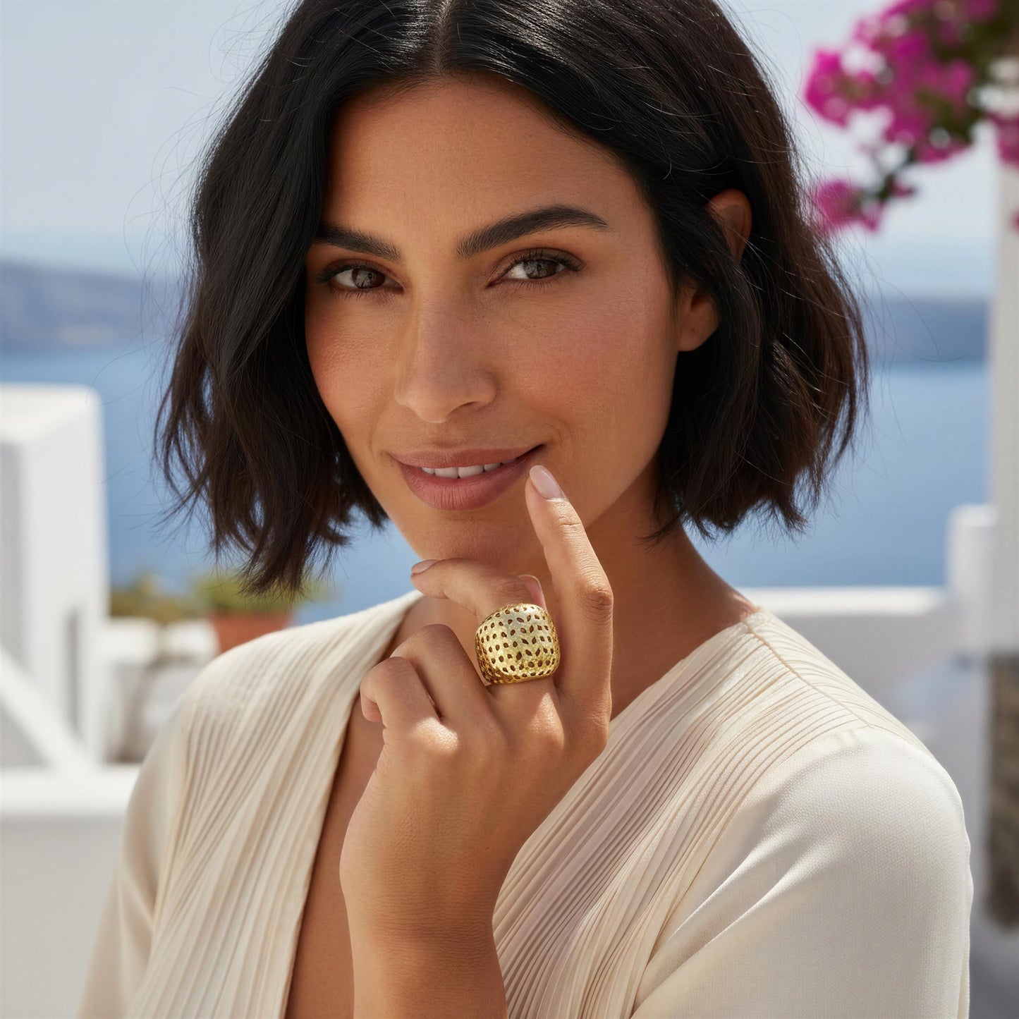A woman with short dark hair wearing a large, chunky gold ring with a textured, honeycomb-like pattern. She is wearing a cream-colored pleated top, with a blurred background showing blue water and white buildings.