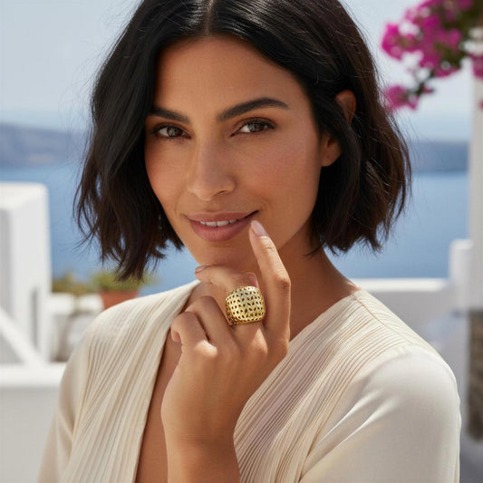 A woman with short dark hair wearing a large, chunky gold ring with a textured, honeycomb-like pattern. She is wearing a cream-colored pleated top, with a blurred background showing blue water and white buildings.