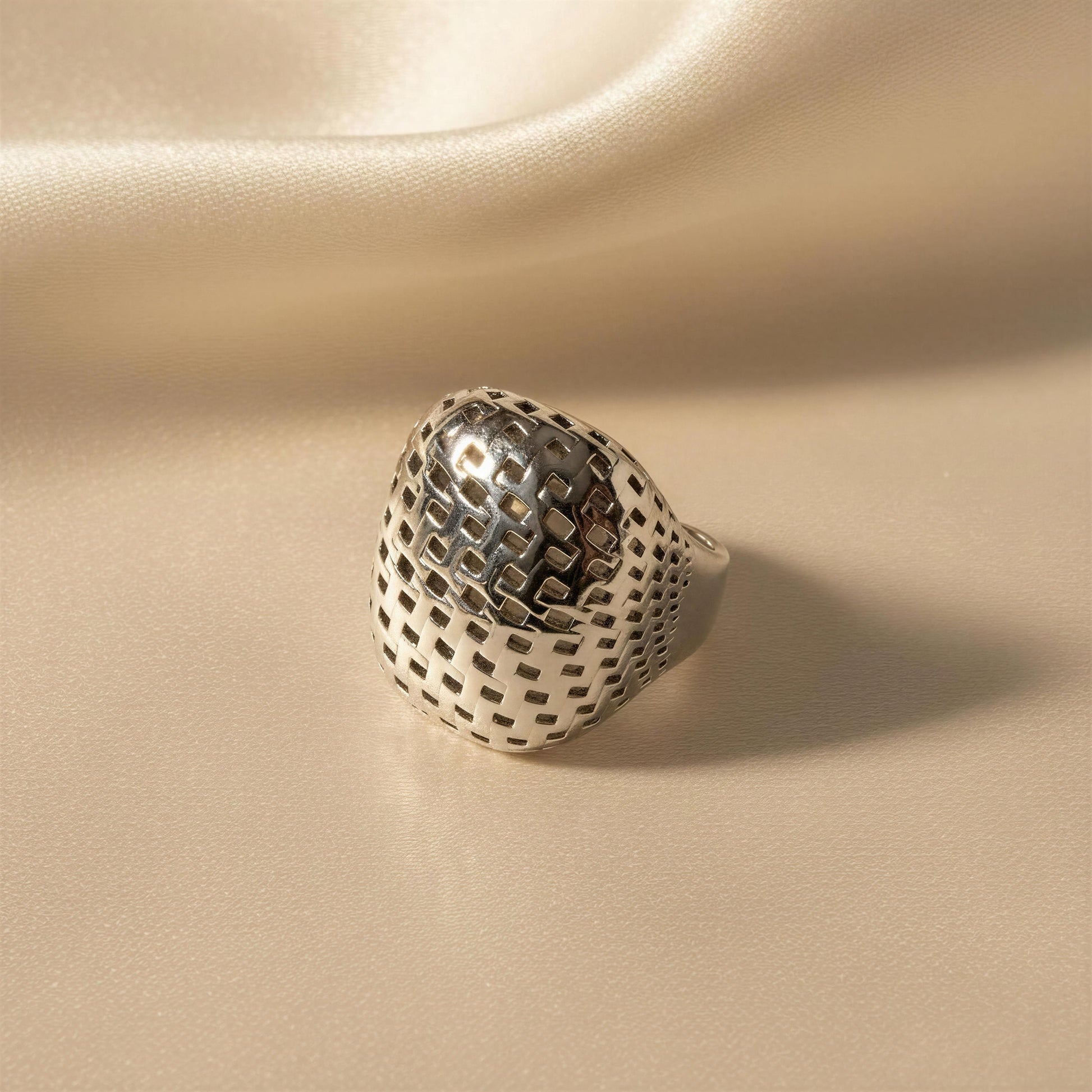 A silver ring with a textured, openwork geometric pattern, resting on a draped beige satin fabric background.