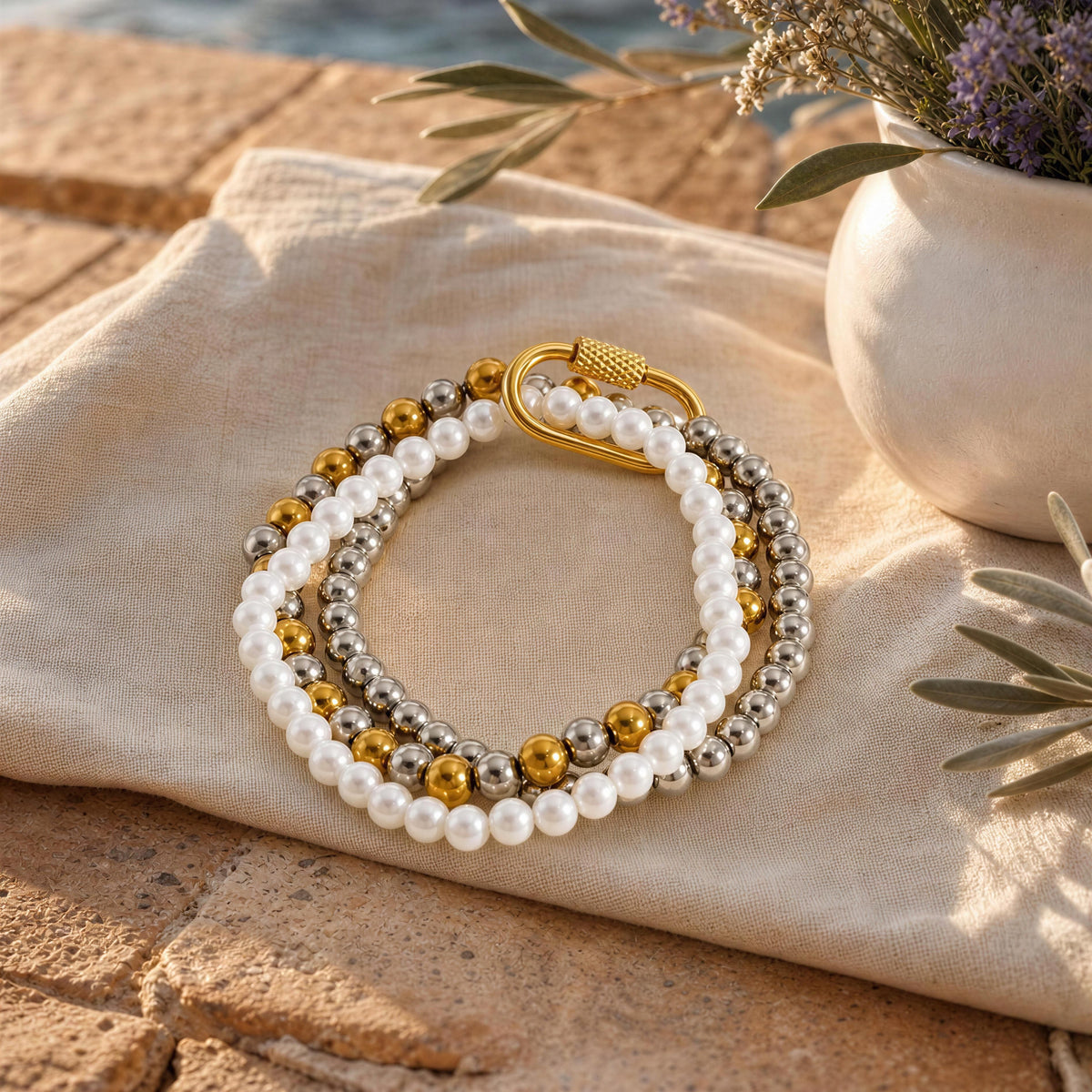 A multi-strand beaded bracelet with white pearls, silver-toned beads, and gold-toned accent beads, displayed on a beige linen cloth outdoors.