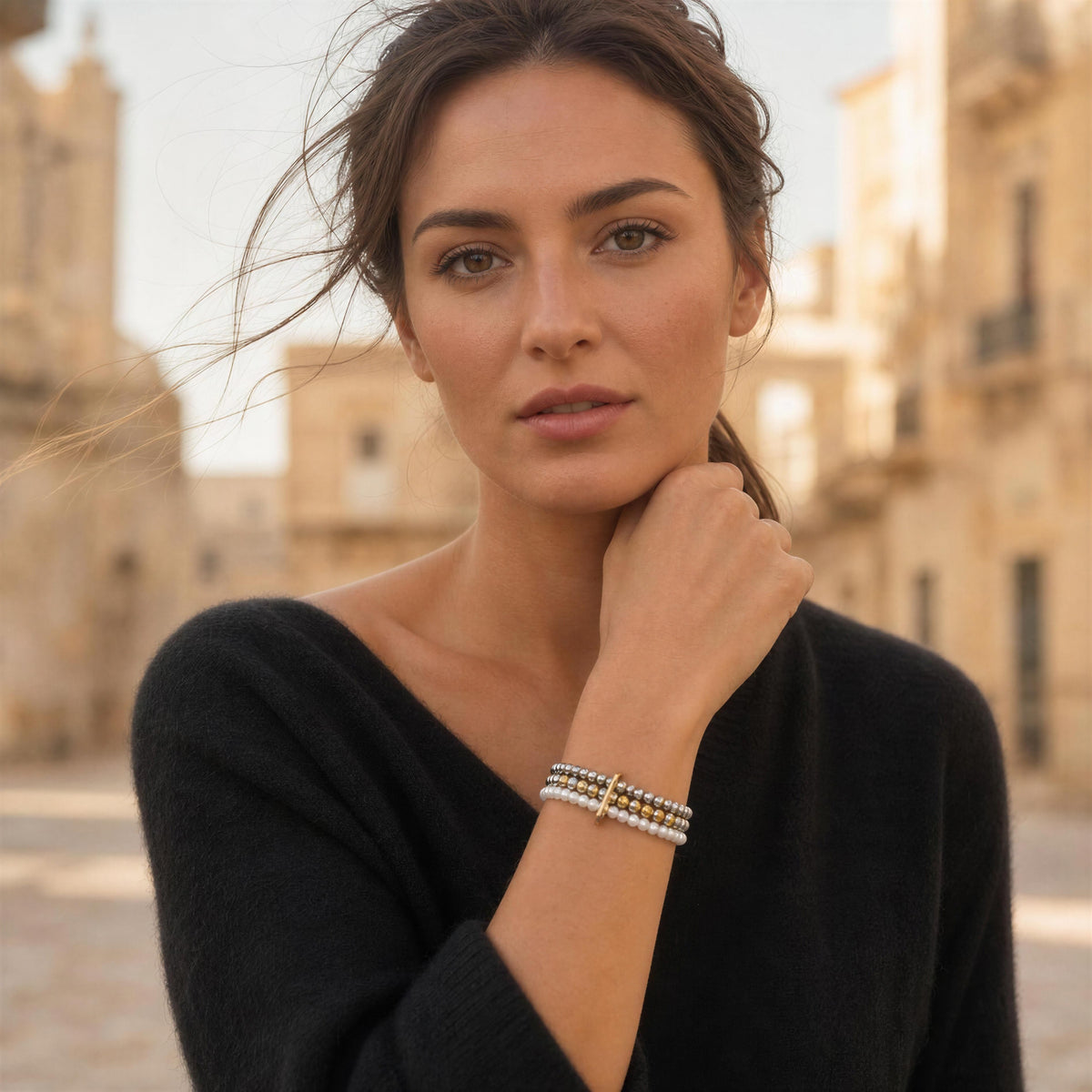 A woman wearing a delicate multi-strand beaded bracelets, posing in an outdoor urban setting with blurred historic buildings in the background.