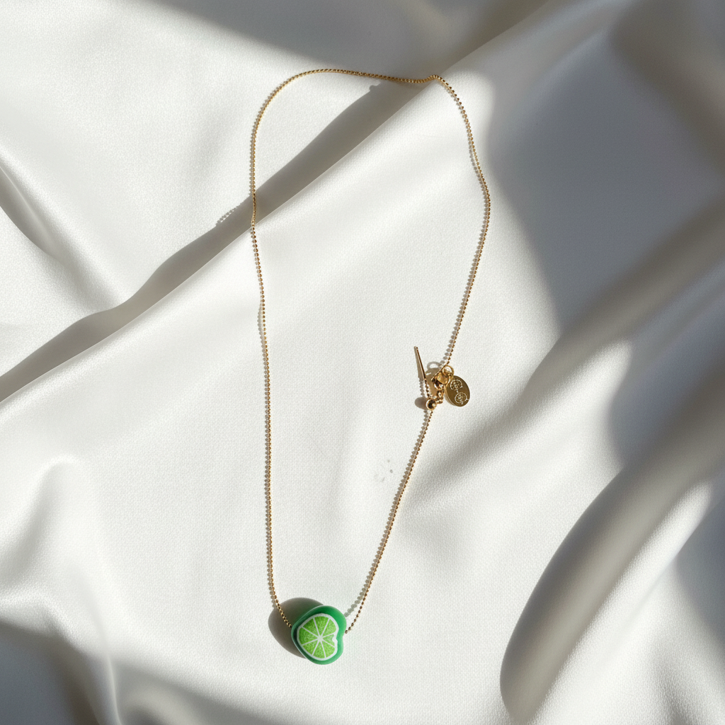 Tutti Frutti Lime Necklace with hand-painted lime charm displayed on white fabric, highlighting the 18K gold-plated adjustable ball chain and sliding bar clasp. 