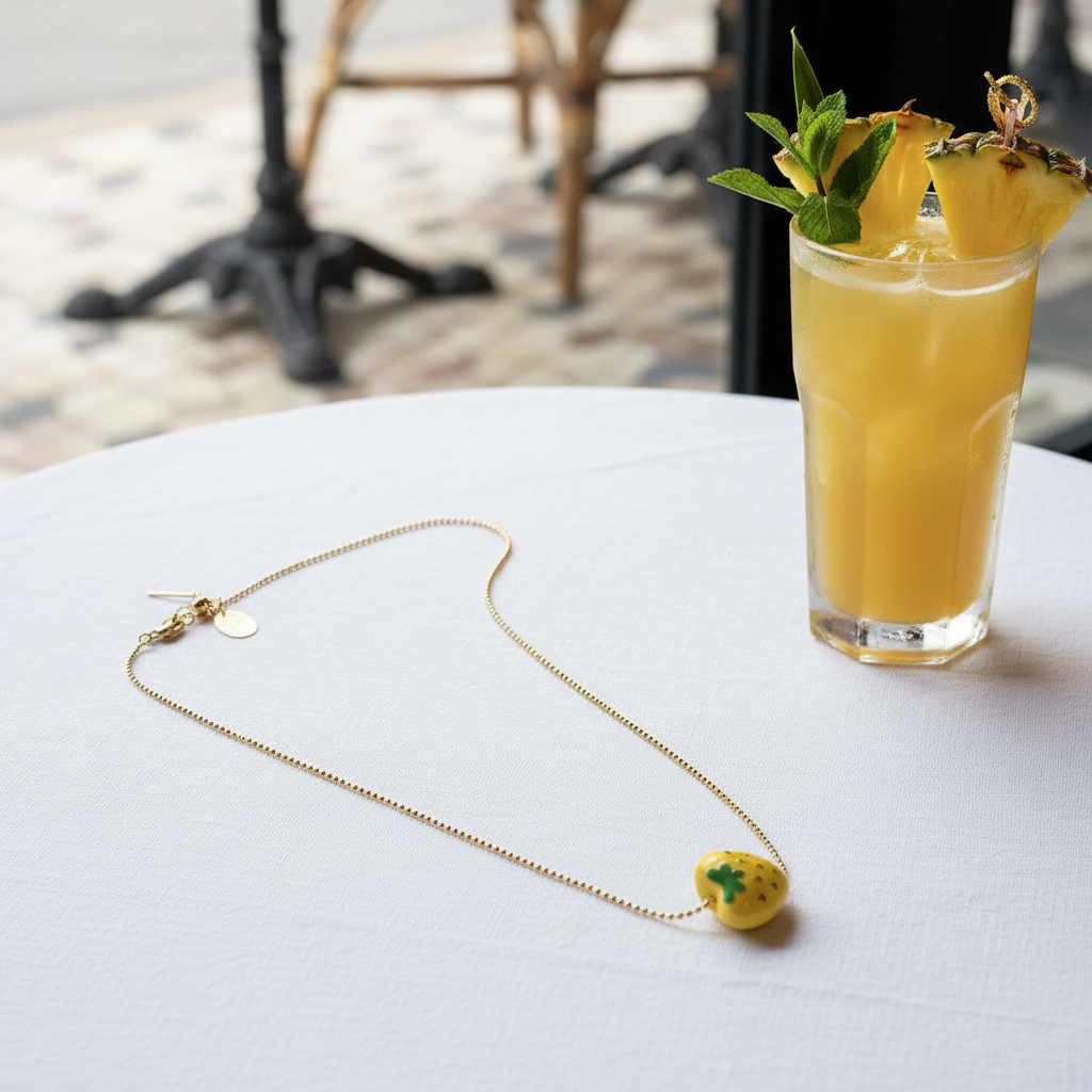 Gold chain pineapple necklace styled beside a yellow cocktail on white café table