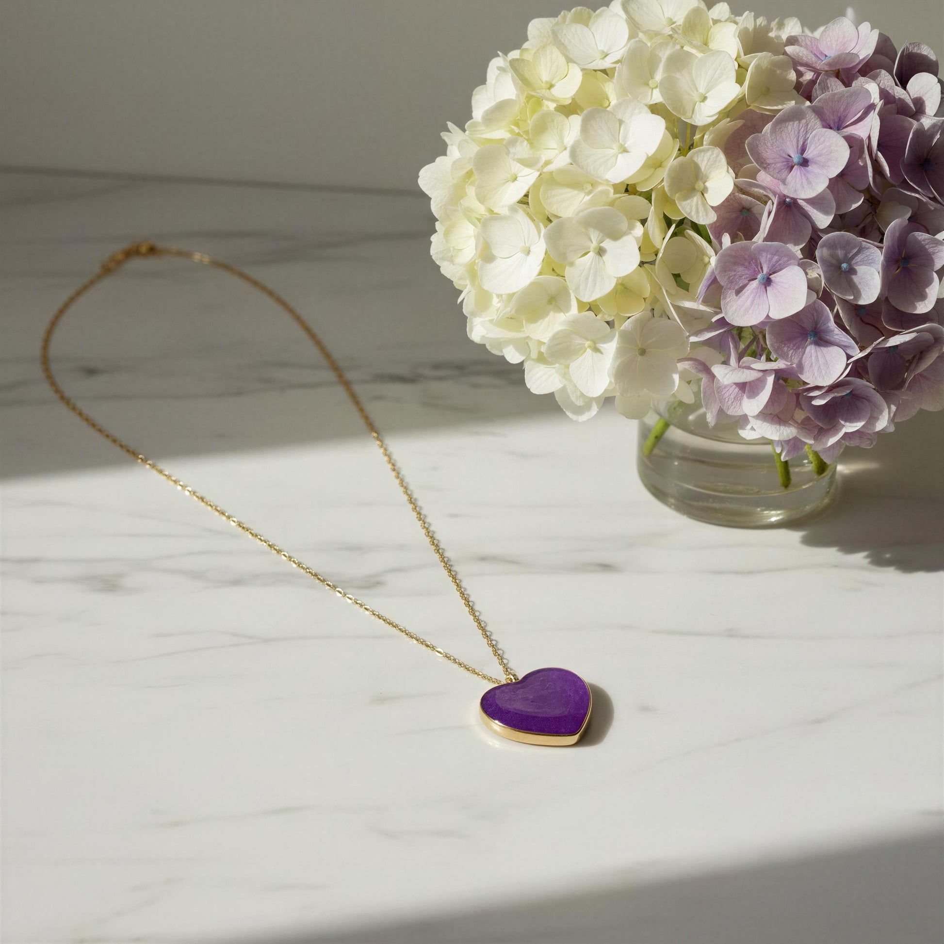 Purple heart-shaped pendant on a gold chain with flowers in the background. The Viola_Heart_Necklace from Chica Chica Jewelry.