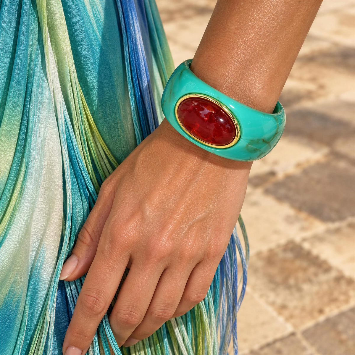 A close-up of a person's hand and wrist wearing the Vittoria Bangle from Chica Chica Jewelry; a chunky turquoise bangle bracelet with a large oval burgundy cabochon and gold trim, set against a background of a colorful striped skirt and stone pavement.