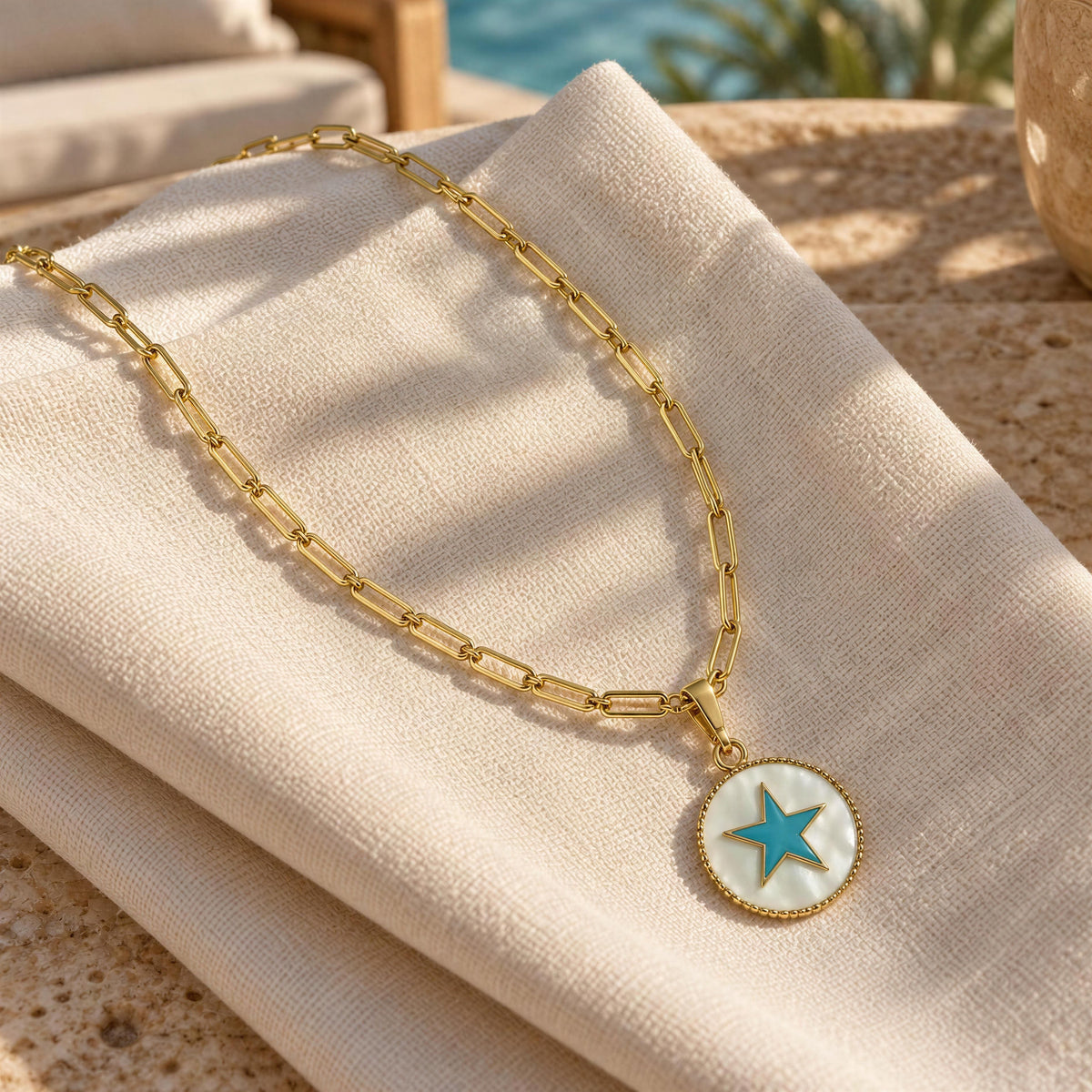 A gold chain necklace with a round pendant featuring a turquoise blue star on a white background, draped over a beige linen cloth in warm sunlight.