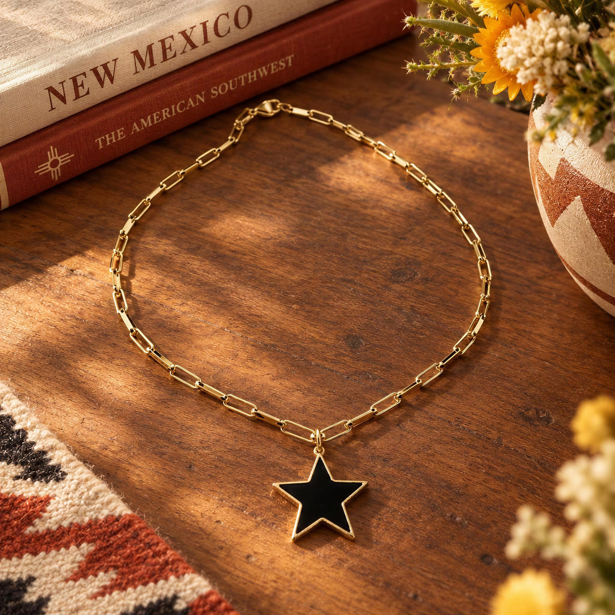 A gold chain necklace with a black star pendant, styled on a wooden tabletop with a book, flowers, and a patterned textile nearby.