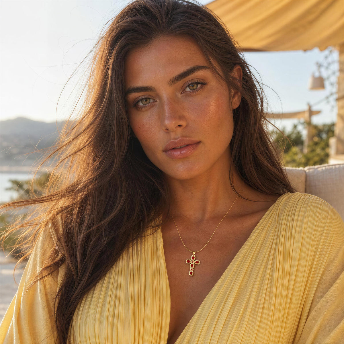 A woman with long brown hair wearing a yellow pleated top and a delicate gold cross pendant necklace, photographed outdoors with a warm, sunny background.