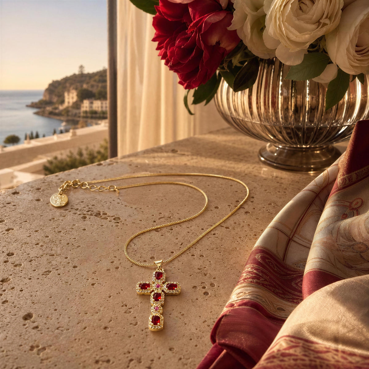 A gold cross pendant necklace with red gemstone accents lies on a stone tabletop beside a vase of flowers and a draped scarf, with a coastal window in the background.